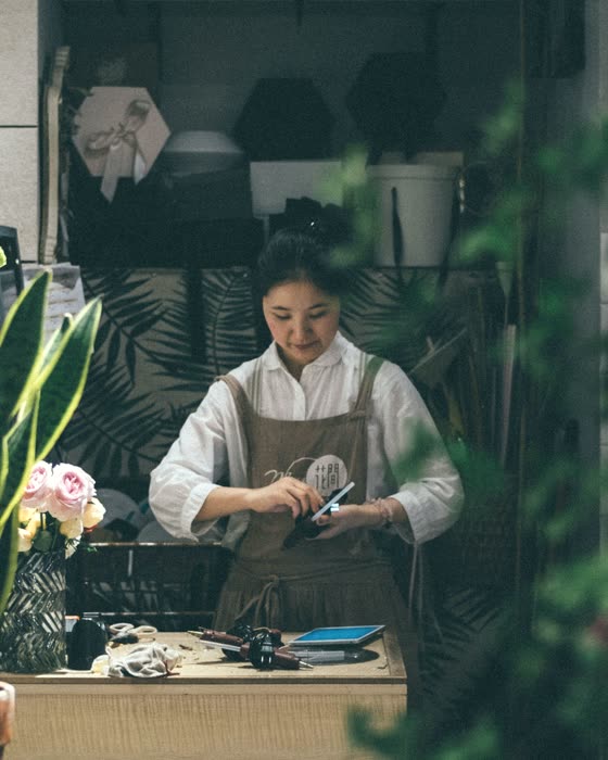 A florist checking her phone at a worktable surrounded by stock.