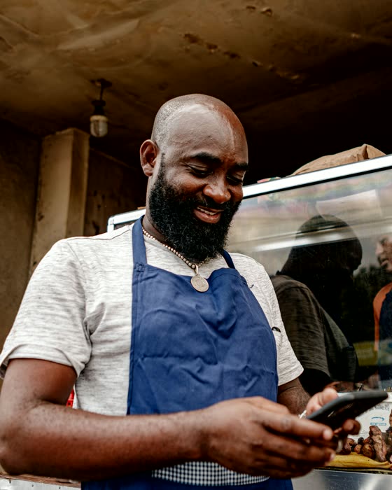 A street food vendor in an apron reading his phone beside a grill case.